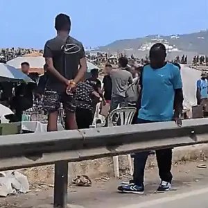 Personas reunidas detrás de una barrera metálica, con montes al fondo