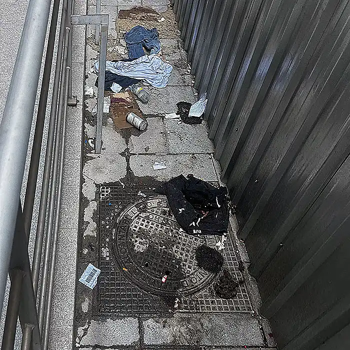 Vista de un pasillo estrecho entre barandilla y pared metálica con ropa y residuos en el suelo