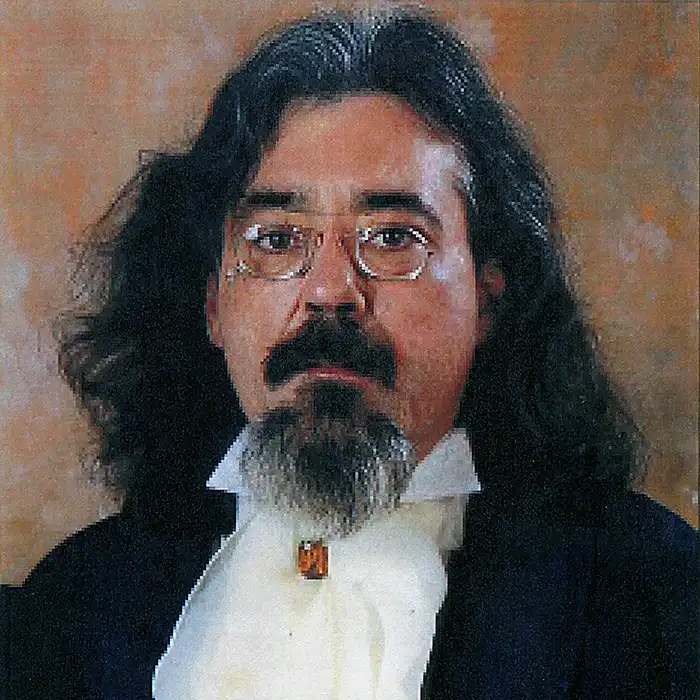 Retrato de una persona con cabello largo oscuro, gafas y barba sobre fondo marrón